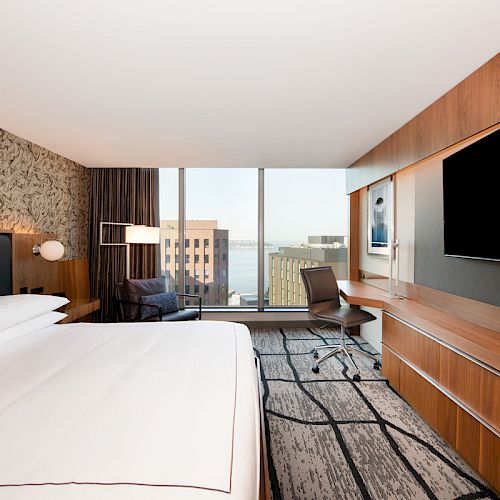 A modern hotel room features a large bed, a desk, an office chair, a flat-screen TV, and a city view through floor-to-ceiling windows.