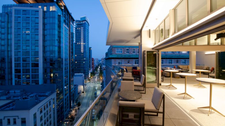 A modern rooftop terrace with seating overlooks a city street at dusk, surrounded by tall buildings with large windows and lights.