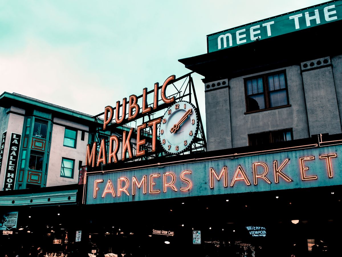 The image shows a notable landmark of a "Public Market" and "Farmers Market" with neon signs, and a clock, against a backdrop of urban buildings.