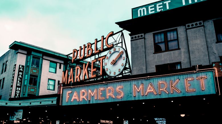 The image shows a notable landmark of a "Public Market" and "Farmers Market" with neon signs, and a clock, against a backdrop of urban buildings.