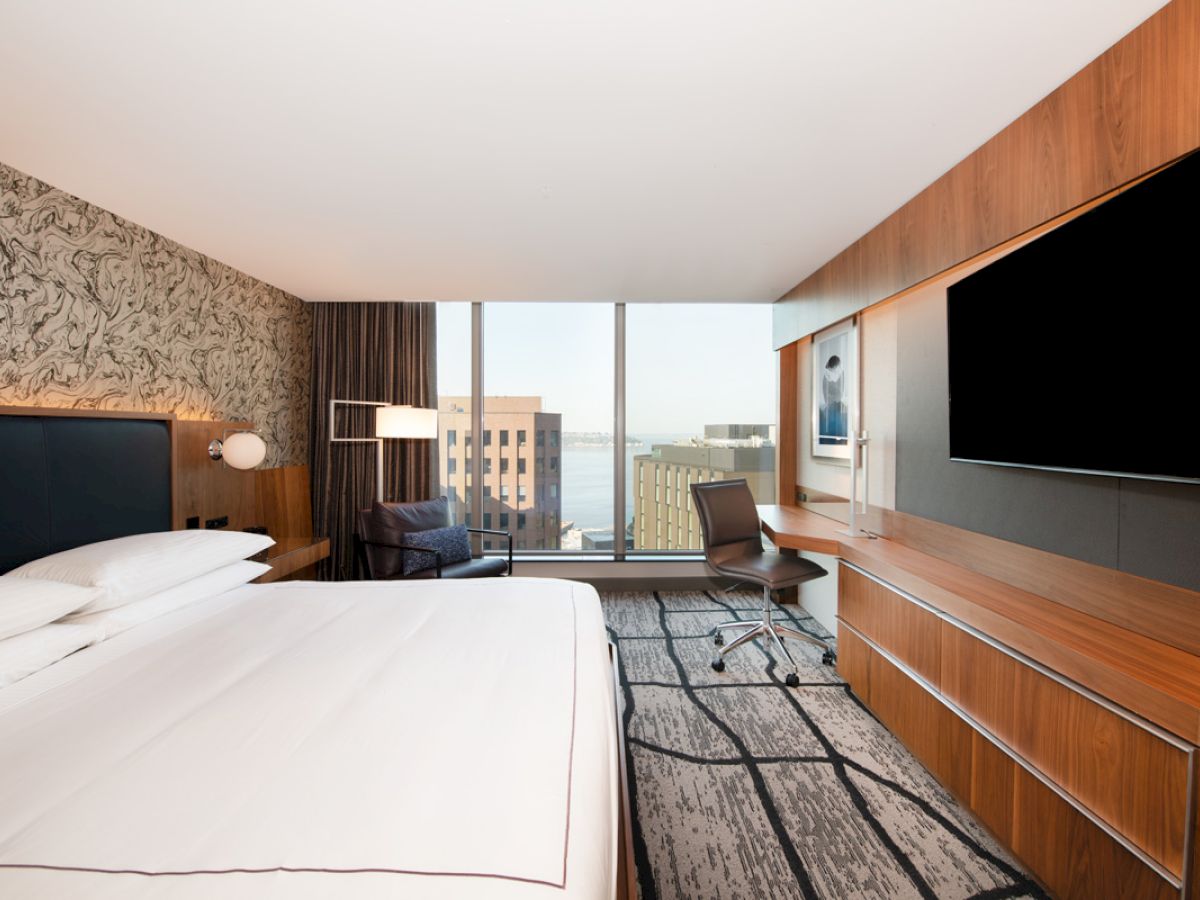 A modern hotel room with a large bed, wall-mounted TV, desk, chair, and window view of buildings and water.