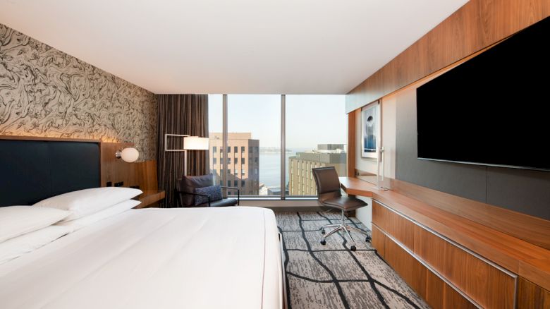 A modern hotel room with a large bed, wall-mounted TV, desk, chair, and window view of buildings and water.