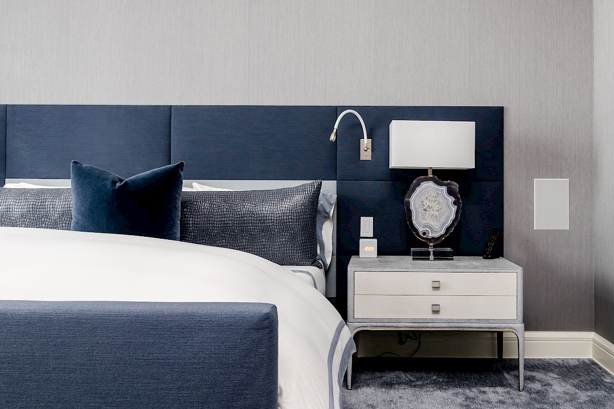 A modern bedroom with a blue upholstered headboard, white bedding, a lamp, and decor on a nightstand, set against a gray wall.