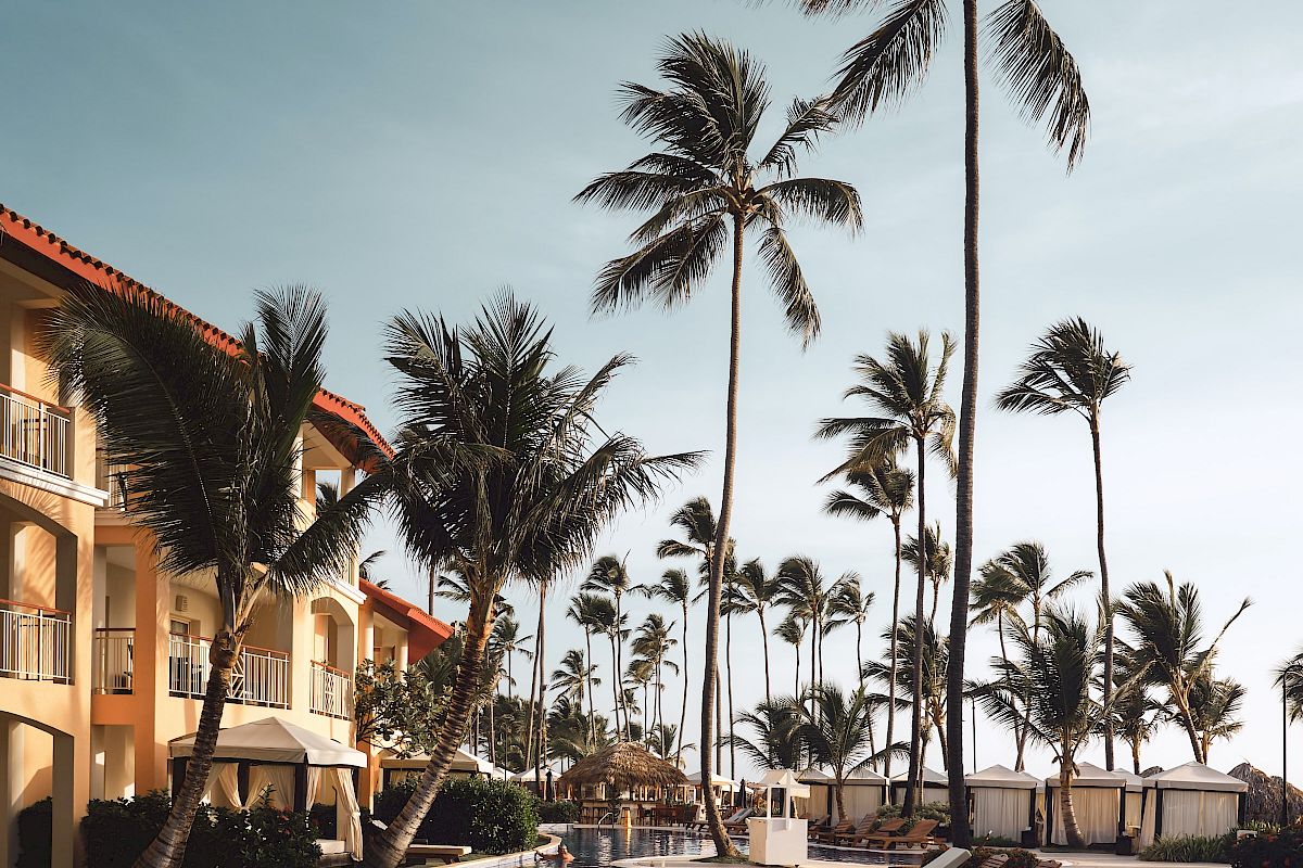 A luxurious poolside scene with tall palm trees, lounge chairs, and a multi-story building in the background, all under a clear blue sky.