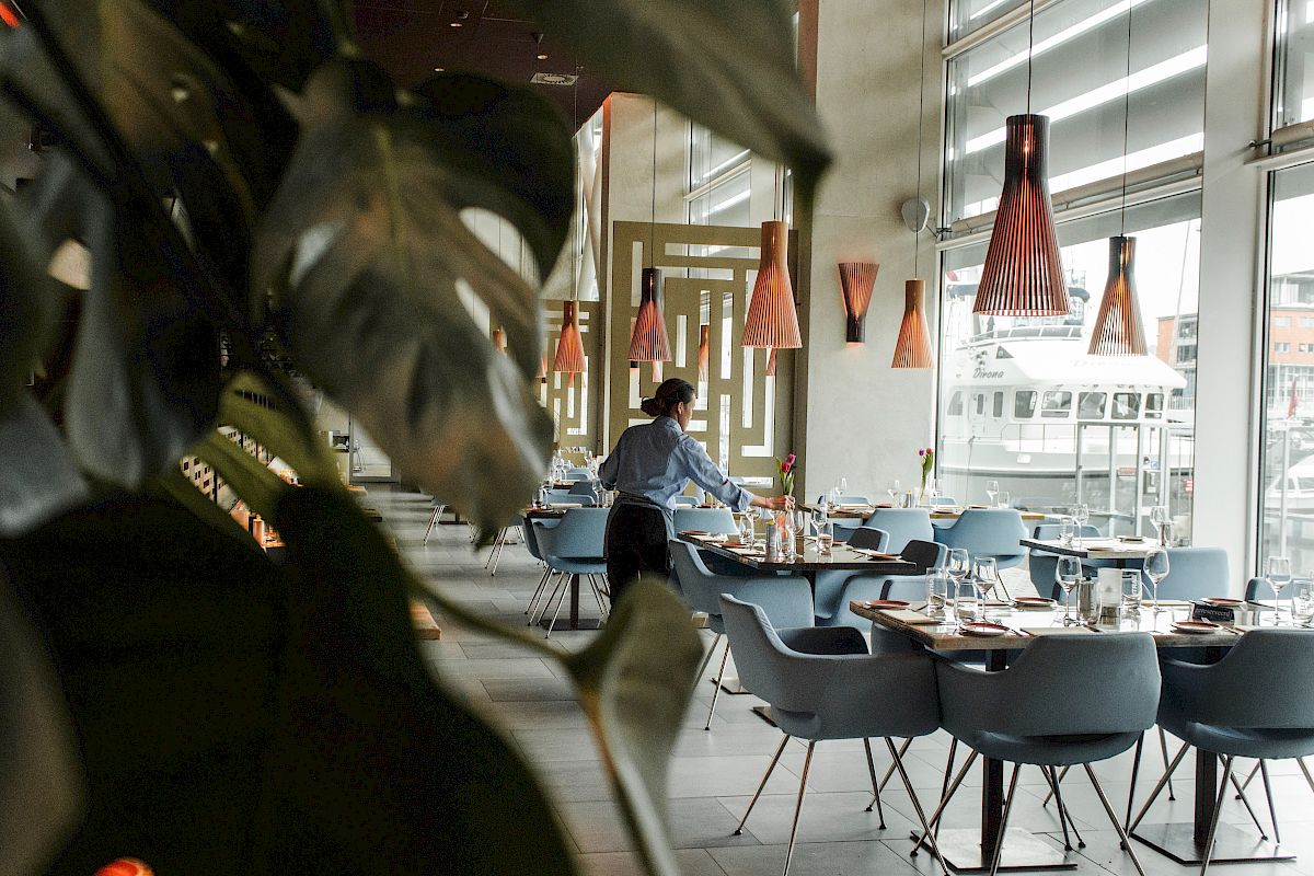 A person is arranging tables in a modern restaurant with large windows, stylish lighting, and gray-blue chairs.