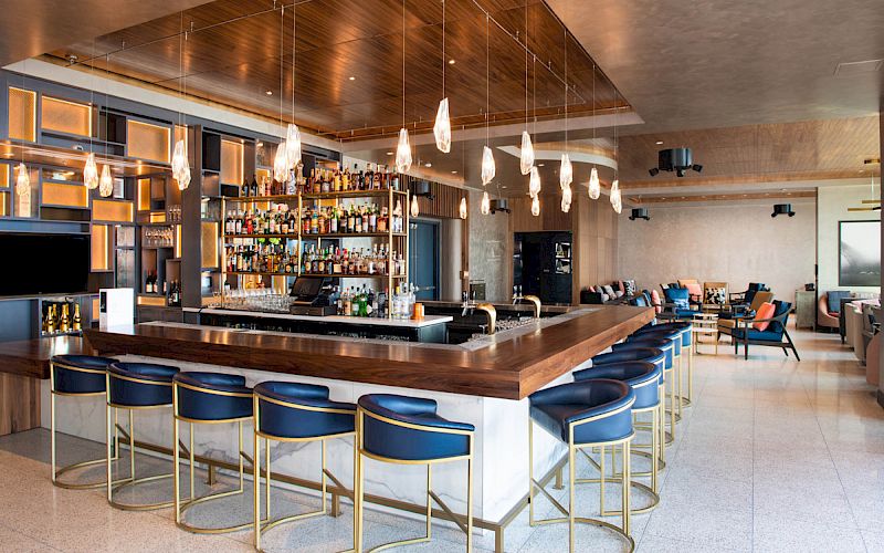 The image shows a modern bar with wooden accents, stylish blue chairs, pendant lighting, and a well-stocked shelf of various bottles.