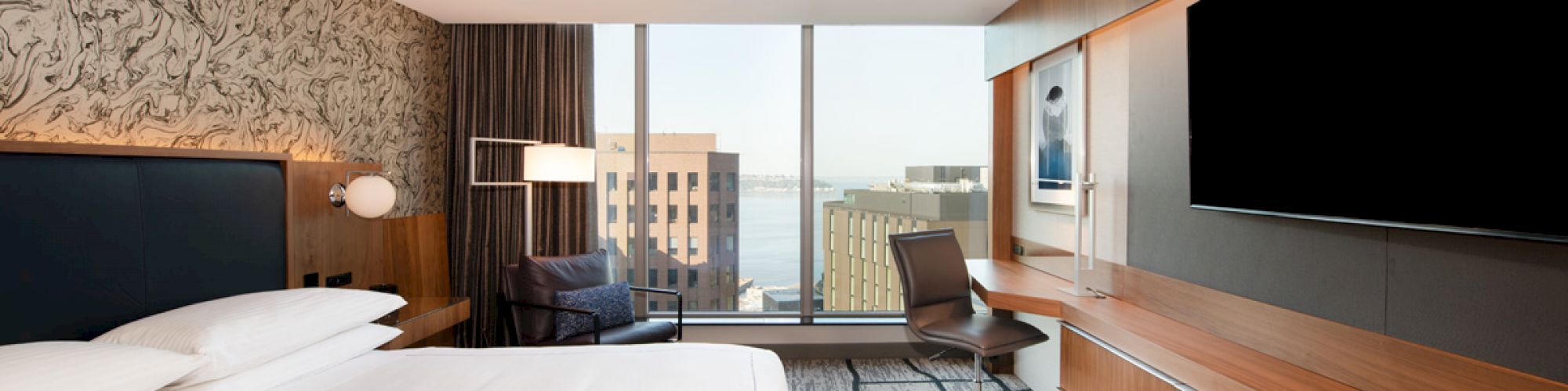 A modern hotel room with a large bed, wall-mounted TV, wooden desk, and a window view of city buildings. There is patterned wallpaper behind the bed.