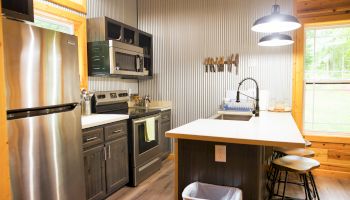 A modern kitchen with stainless steel appliances, wooden cabinets, a countertop island with stools, and a window letting in natural light.