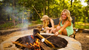 Two people are sitting around an outdoor fire pit, roasting marshmallows on sticks, surrounded by a forested area and enjoying the campfire.