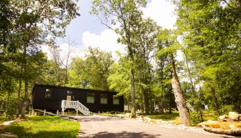 The image shows a small, dark-colored cabin with white steps, surrounded by trees and located in a forested area on a sunny day.