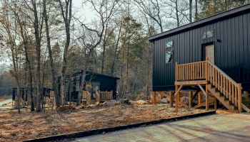 The image shows modern tiny houses with wooden stairs set among trees in a forested area, surrounded by pathways and natural vegetation.