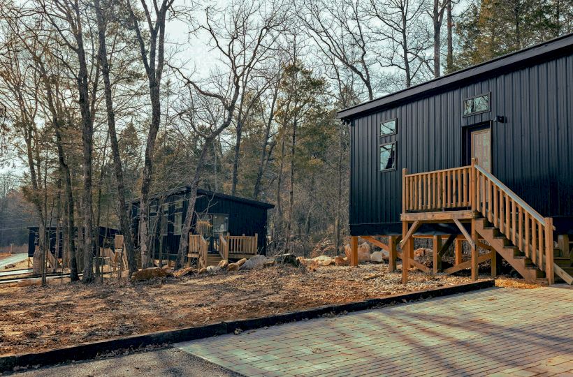 The image shows modern tiny houses with wooden stairs set among trees in a forested area, surrounded by pathways and natural vegetation.