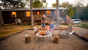 A family gathers around a fire pit in front of a cabin, roasting marshmallows, with a white van parked nearby and trees in the background.