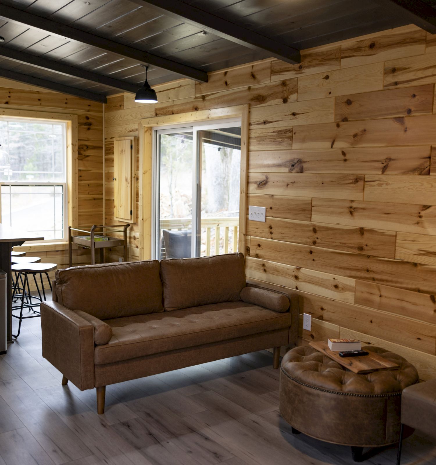 This image shows a cozy living room and kitchen with wood-paneled walls, a brown leather couch, a round ottoman, and stainless steel kitchen appliances.