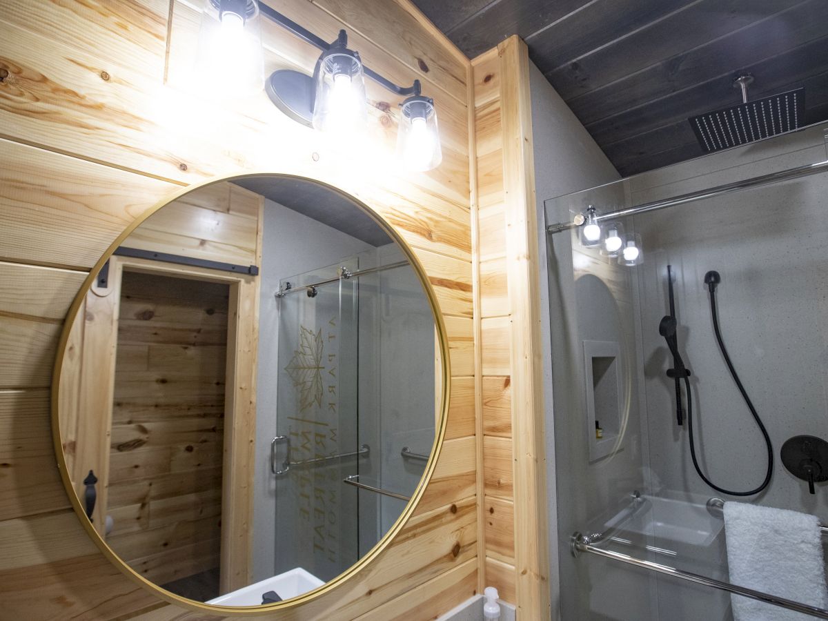 A modern bathroom features a round mirror with wood paneling, a glass-enclosed shower, a black showerhead, and illuminated lighting fixtures.