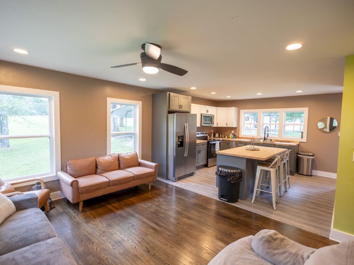 A cozy open-plan living room and kitchen with a ceiling fan, sofas, a kitchen island, and modern appliances, featuring wood flooring.