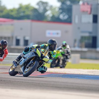 Three motorcyclists are racing on a track, leaning into a turn, with a blurred building and trees in the background.