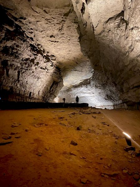 A large, illuminated cave with a rough rocky floor and silhouetted figures in the distance near the cave's wall.